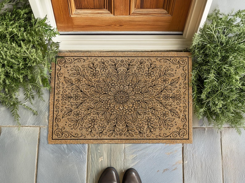 A top-down view of a rectangular tan coir doormat featuring an elaborate black botanical mandala design. The pattern radiates from a central flower, surrounded by layers of detailed petals, leaves, and smaller blossoms that fill the entire mat. Decorative scrollwork flourishes accent the four corners, all enclosed by a thin black rectangular border.