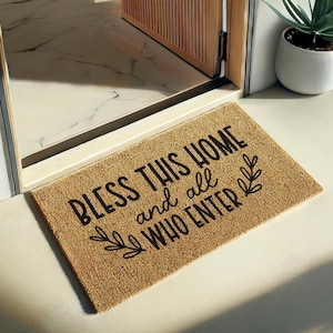 Può includere: Un zerbino rettangolare in fibra naturale con la scritta nera "BLESS THIS HOME and all WHO ENTER". Il tappeto ha un design a foglie ed è posizionato davanti a una porta.