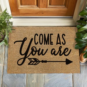 Peut inclure: Un paillasson rectangulaire en fibres naturelles, avec l'inscription "COME AS You are" en noir et un motif de flèche. Le tapis est placé devant une porte en bois.