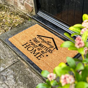 Può includere: Un zerbino rettangolare in fibra di cocco naturale. Il tappeto ha un bordo nero e presenta la frase "Everything that matters most is at HOME" in nero, con un disegno di una casa. Posizionato davanti a una porta nera.