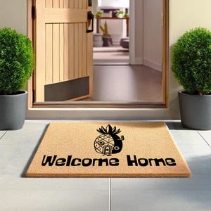 Op de afbeelding: Een rechthoekige deurmat met de woorden "Welcome Home" in zwarte tekst. Een zwart ananasontwerp staat boven de tekst. De mat is bruin en ligt voor een houten deur.