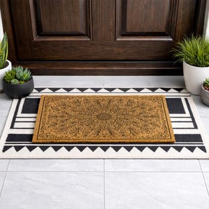 K&ouml;nnte beinhalten: Eine rechteckige Kokos-Fu&szlig;matte mit detailliertem floralem Design liegt auf einem schwarz-wei&szlig; gemusterten Teppich. Die Szene spielt sich vor einer dunkelbraunen T&uuml;r ab, flankiert von Topfpflanzen, die einen einladenden Eingang schaffen.