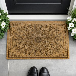 K&ouml;nnte beinhalten: Eine rechteckige Fu&szlig;matte mit einem detaillierten schwarzen Blumen-Mandala-Design auf einem nat&uuml;rlichen, hellbraunen Hintergrund. Die Matte liegt vor einer schwarzen T&uuml;r, flankiert von Topfpflanzen mit wei&szlig;en Bl&uuml;ten. Schwarze Schuhe sind unten zu sehen.