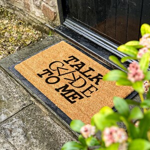 May include: A rectangular coir doormat with the words "TALK TO ME" in black capital letters. The mat is placed on a stone step near a black door. The coir mat is made of natural fibres.