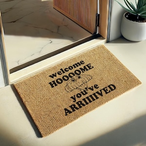 May include: A rectangular doormat made of natural fibers, featuring the text "welcome HOOOOOME you've ARRIVED" in black. A graphic of a dog's face is in the center. The mat is placed in a doorway.