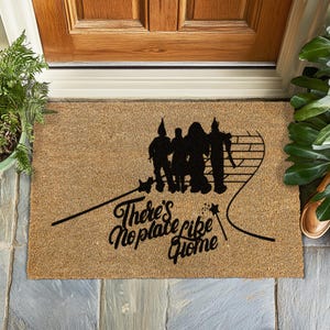 May include: A brown coir doormat with the text "There's No Place Like Home" and a black silhouette of the Wizard of Oz characters walking down the yellow brick road.