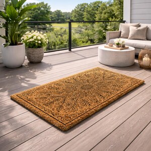 K&ouml;nnte beinhalten: Rechteckige Fu&szlig;matte mit detailliertem floralem Design in Schwarz auf tanfarbenem Hintergrund. Die Matte besteht aus Naturfasermaterial und liegt auf einer Holzterrasse. Die Szene umfasst Topfpflanzen, ein Sofa und einen runden Tisch.