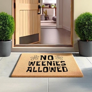 May include: A rectangular doormat made of natural fibers with the text "NO WEENIES ALLOWED" in black. The mat is placed in front of a wooden door, with two potted green plants on either side.