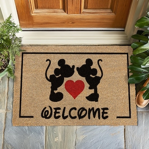 Peut inclure: Un paillasson en fibre de coco brun avec une silhouette noire de Mickey Mouse et Minnie Mouse s'embrassant avec un cœur rouge entre eux. Le texte "WELCOME" est écrit en cursive noire en dessous des personnages.