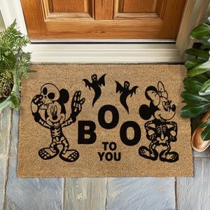 Peut inclure: Un paillasson rectangulaire en fibre de coco sur le thème d'Halloween. Le tapis présente des silhouettes noires de Mickey et Minnie Mouse en forme de squelette, ainsi que trois fantômes et la phrase "BOO TO YOU."