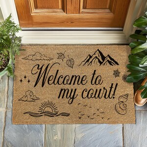 May include: A rectangular doormat made of natural fibers. The mat features the phrase "Welcome to my court!" in elegant script, along with mountain, cloud, leaf, and sun illustrations. The design is in black against a tan background.