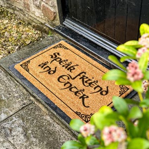 May include: A rectangular doormat made of natural coir fiber. The mat features the words "Speak Friend and Enter" in a decorative black font. The mat is framed with a black border and sits on a stone surface near a black door.