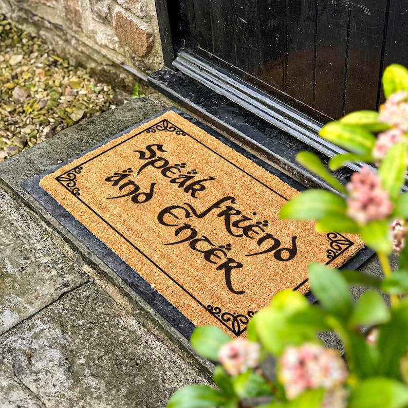 May include: A rectangular doormat made of natural coir fiber. The mat features the words "Speak Friend and Enter" in a decorative black font. The mat is framed with a black border and sits on a stone surface near a black door.