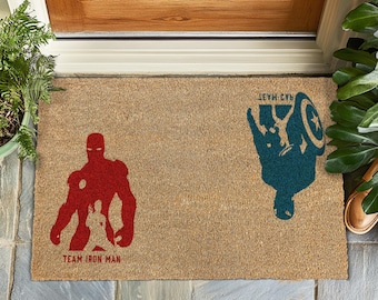 Könnte beinhalten: Eine braune Kokosfaser-Fußmatte mit dem Text "Team Iron Man" und einer roten Silhouette von Iron Man auf der linken Seite. Auf der rechten Seite steht der Text "Team Cap" und eine blaue Silhouette von Captain America.