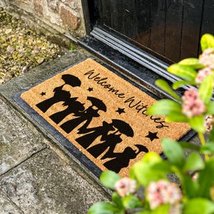Peut inclure: Un paillasson rectangulaire en fibre de coco avec une bordure noire. Le tapis présente les mots "Welcome Witches" en écriture cursive, ainsi que des silhouettes noires de sorcières et d'étoiles. Le tapis est posé sur une surface en pierre.