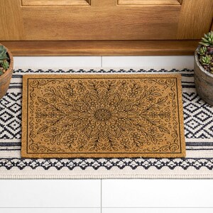 K&ouml;nnte beinhalten: Eine rechteckige Fu&szlig;matte mit floralem Design in der Mitte, platziert auf einem schwarz-wei&szlig; gemusterten Teppich. Die Fu&szlig;matte besteht aus einem hellbraunen Material. Zwei Topf-Sukkulenten flankieren den Teppich in der N&auml;he einer Holzt&uuml;r.
