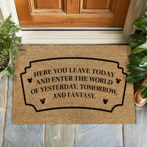 Peut inclure: Un paillasson en fibre de coco brun avec le texte "HERE YOU LEAVE TODAY AND ENTER THE WORLD OF YESTERDAY, TOMORROW, AND FANTASY." en lettres noires. Le texte est encadré d'une bordure noire avec des oreilles de Mickey Mouse à chaque extrémité.