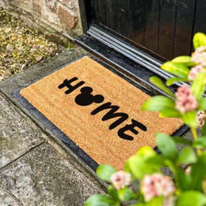 May include: A rectangular coir doormat with the word "HOME" in black, with the "O" replaced by a Mickey Mouse head silhouette. The mat is placed in front of a black door. The mat is made of natural coir fiber.