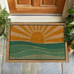 Könnte beinhalten: Eine braune Kokosfaser-Fußmatte mit einem grafischen Design einer gelben Sonne, die über grünen Wellen aufgeht. Die Sonne hat Strahlen, die von ihr ausgehen.