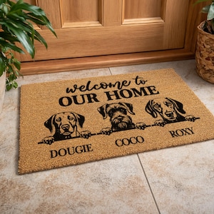 May include: A rectangular doormat with the words "welcome to OUR HOME" in black. Below the text are three dog illustrations with the names "DOUGIE", "COCO", and "ROXY" printed below each dog. The mat is made of a natural fiber material.