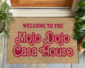 May include: A rectangular doormat made of natural fibers. The mat features the text "WELCOME TO THE Mojo Dojo Casa House" in a vibrant red color. The mat is placed in front of a wooden door.