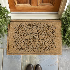 May include: A rectangular doormat made of natural coir fiber, featuring a symmetrical floral design in black. The design includes a large central flower surrounded by leaves and smaller flowers. The mat is placed in front of a wooden door.