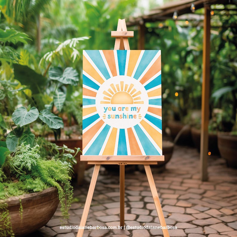May include: A watercolor painting on canvas with a sunburst design in shades of blue, orange, and yellow. The sunburst design surrounds a stylized sun with the text "you are my sunshine" in the center.