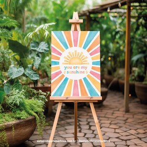 May include: A watercolor painting on canvas with a sunburst design in shades of orange, yellow, pink, and blue. The sunburst design has a sun in the center with the text "You are my sunshine" written below it.