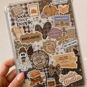 以下が含まれることがあります： カボチャ、松ぼっくり、お化け、そして「Books lover people」、「Books+Coffee」、「Official Book Club Member」、「Sorry I was reading」、「Sweater Weather」などのテキストが描かれた、秋をテーマにしたステッカーで覆われた透明なタブレット保護ケース。