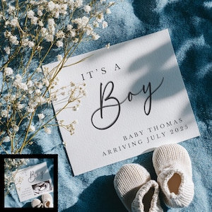 May include: A white card with the text "IT'S A Boy" and baby announcement details, accompanied by a pair of beige baby booties. The card also includes the text "BABY THOMAS ARRIVING JULY 2025". The background is a blue fabric with white flowers.