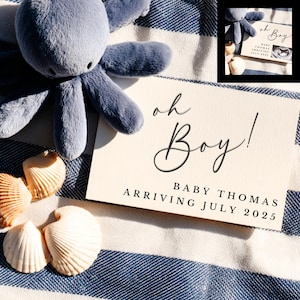Op de afbeelding: Een zacht, blauw pluche octopus speelgoed ligt naast een crème kaart met de tekst "oh Boy! BABY THOMAS ARRIVING JULY 2025." Schelpen zijn gerangschikt op een blauw-wit gestreepte stof.