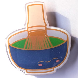 May include: A blue and white ceramic bowl with a green liquid and a brown whisk inside. The bowl has a cute cartoon face with pink cheeks and a smile.