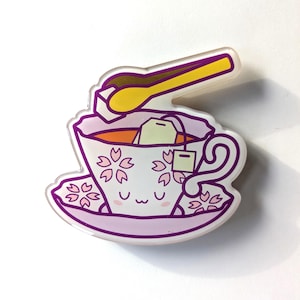 May include: A purple teacup with a pink floral pattern and a cute face. The teacup is filled with tea and a tea bag. A yellow sugar spoon is placed on top of the teacup.