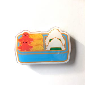 May include: A clear acrylic pin featuring a cartoon bento box design. The box is blue with orange trim and contains orange octopus characters, yellow rolled omelets, and white rice balls with green accents.