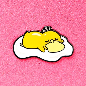 May include: A yellow cartoon duck pin with a large yellow beak and closed eyes, resting on a white egg-shaped surface with a black outline. The pin is set against a pink background.