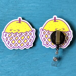 May include: Two badge reels with a yellow fruit design. The fruit has a smiling face and is surrounded by a purple and white basket design. One reel has a black retractable clip and a clear plastic strap.