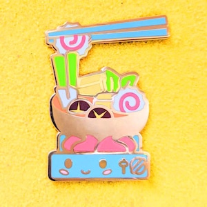 May include: A gold-colored enamel pin depicting a bowl of ramen with chopsticks, noodles, and various toppings. The bowl is sitting on a blue base with a smiling face and a chopstick symbol.