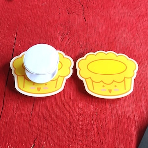 May include: Two yellow phone grips shaped like cartoon egg tarts with scalloped edges, a simple face with two eyes and a small mouth, and a white circular top on one of the grips. The design features a bright yellow center and a lighter yellow crust.