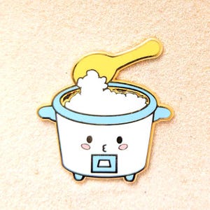 May include: A gold-colored enamel pin of a white rice cooker with a blue lid and a cute cartoon face. The rice cooker has a small blue square on the front and a yellow spoon is sticking out of the top with white rice inside.