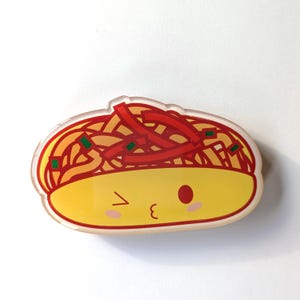 May include: An acrylic pin featuring a cartoon-style bowl of noodles with a yellow face, a winking eye, a closed eye, and a small mouth. The top section displays red noodle strands and small green square garnishes against a light background.