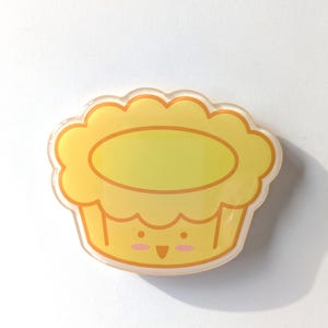 May include: A yellow acrylic pin shaped like a scalloped tart or cupcake. The design features a light yellow top with a darker yellow outline, and a base with a simple face consisting of two small dots for eyes, a small triangle mouth, and pink cheeks.