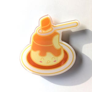 May include: A yellow and orange cartoon pudding with a happy face, topped with caramel sauce and a spoon. The pudding is on a white plate with a yellow rim.