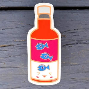May include: A sticker of a bottle with a red top and body, a pink label with three blue fish, and a white bottom with a smiling face. The sticker has a white border and is on a dark gray surface.