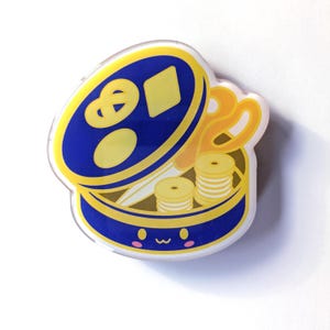 May include: A blue and yellow sewing kit pin with a smiling face. The pin features a blue container with yellow scissors, spools of thread, and a pretzel-shaped design on the lid.