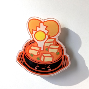 May include: A cartoon illustration of a black pot with a happy face, filled with orange soup and tofu cubes. A cracked orange egg with a yellow yolk sits on top of the soup.