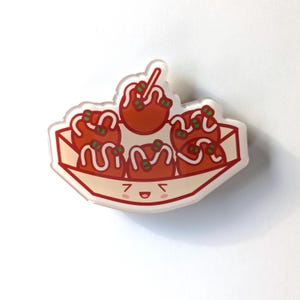 May include: A clear acrylic pin shaped like a takeout container with a smiling face. Inside the container are six red takoyaki balls with green onions and white sauce.
