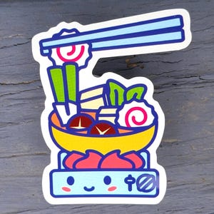 May include: A colorful sticker featuring a cartoon illustration of a bowl of ramen with chopsticks. The bowl is yellow and filled with various ingredients, including mushrooms, vegetables, and a swirl of pink and white fish cake. The sticker is on a gray surface.