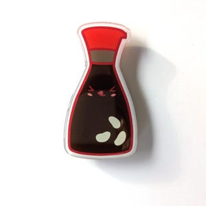 May include: A clear, glossy sticker in the shape of a soy sauce bottle. The bottle has a red cap, brown neck, and a dark brown body with three white spots. The bottle has a smiling face.