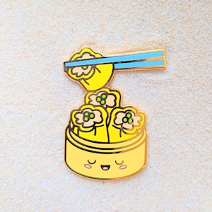 May include: A yellow enamel pin of a cartoon dim sum steamer with three dumplings inside. The steamer has a smiling face and chopsticks are sticking out of the top with a dumpling on the end.