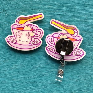 May include: Two badge reels featuring a light pink teacup illustration with a smiling face, cherry blossom patterns, a tea bag, and a yellow spoon. One reel shows the front design, while the other displays the black circular clip mechanism on the back.